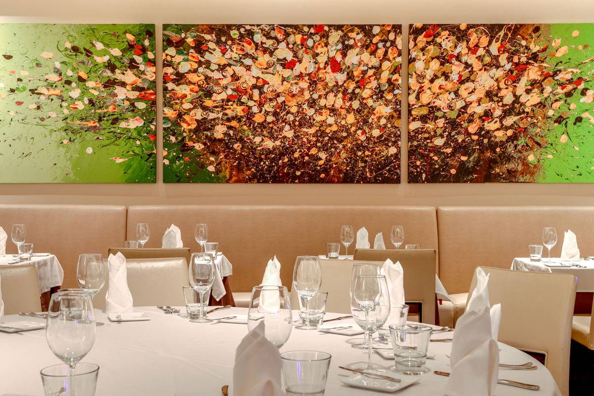 A restaurant interior features white-clothed tables with wine glasses and a large floral mural above a long tan banquette.
