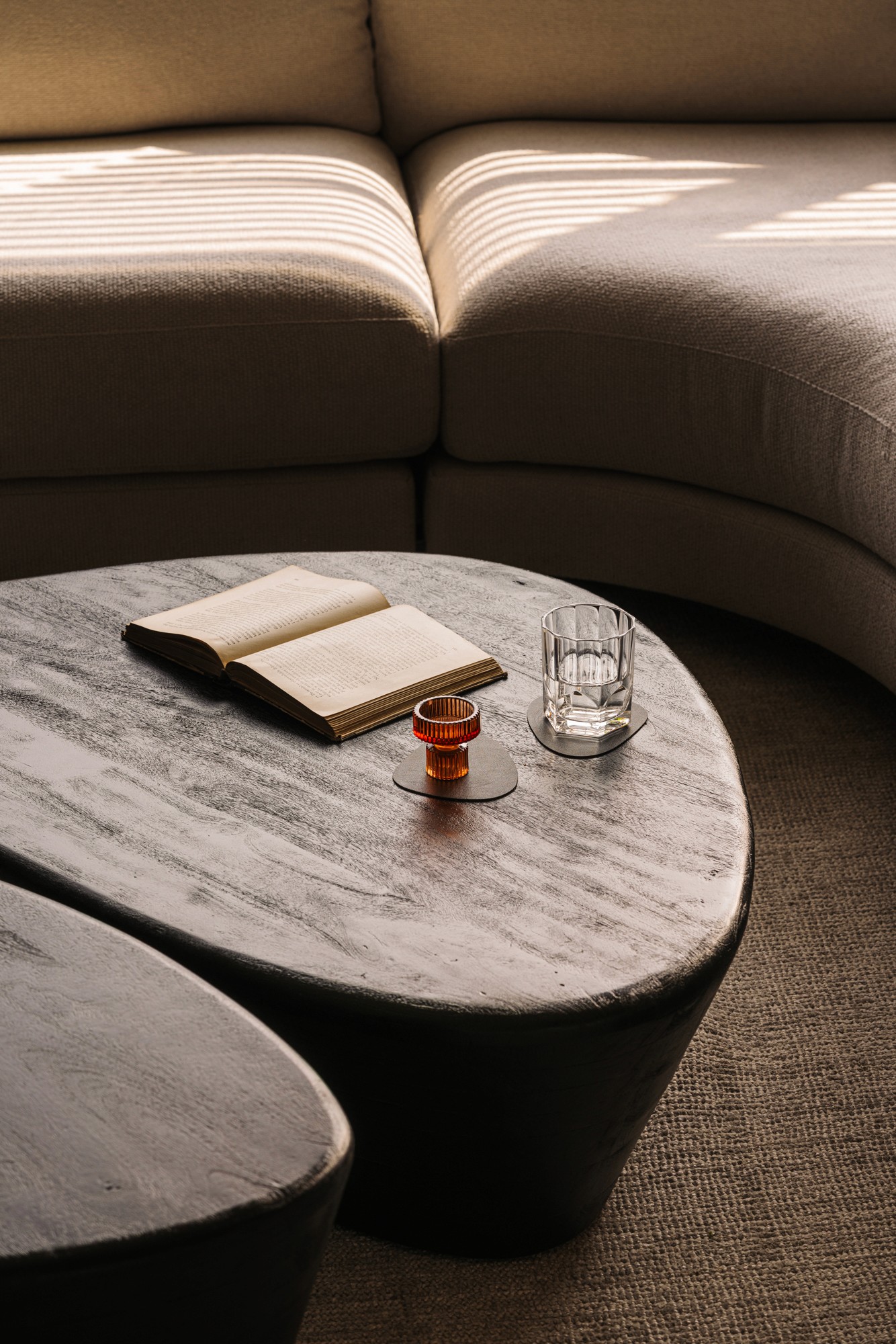 Curved sofa with a sculptural stone coffee table holding an open book and a glass, on a textured rug in soft natural light