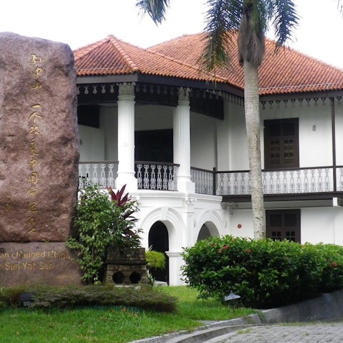 White colonial-style building with red-tiled roof, palm tree, and stone monument inscribed with text, in a landscaped garden.