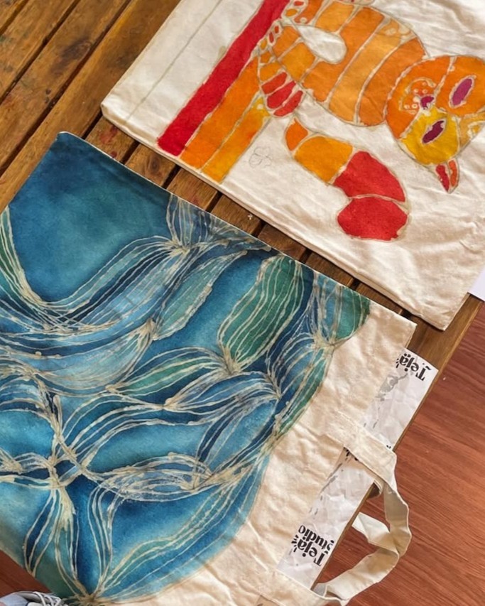 Two colorful tote bags on a wooden surface. The top bag features an orange and red illustration of a playful cat, while the bottom bag showcases a blue and green abstract design resembling waves or plants.