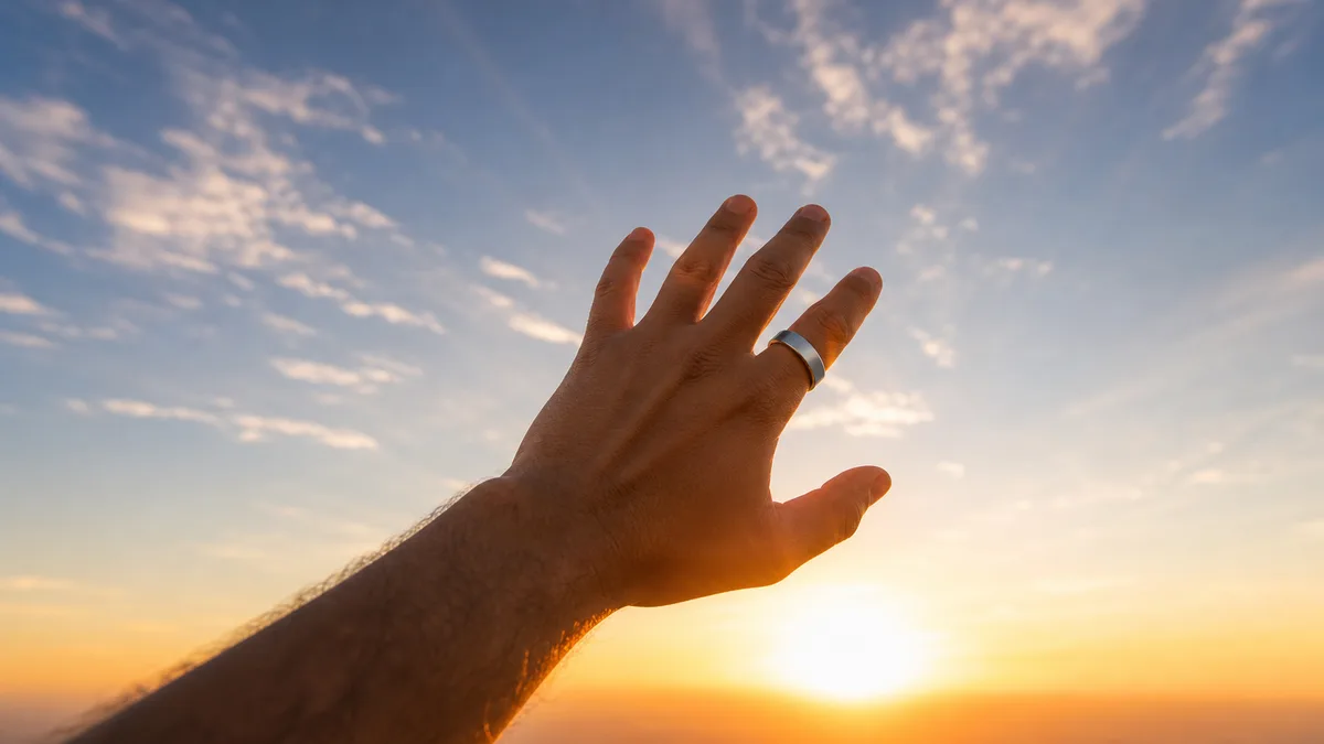 Hand wearing a smart tasbih ring raised toward a golden sunset sky