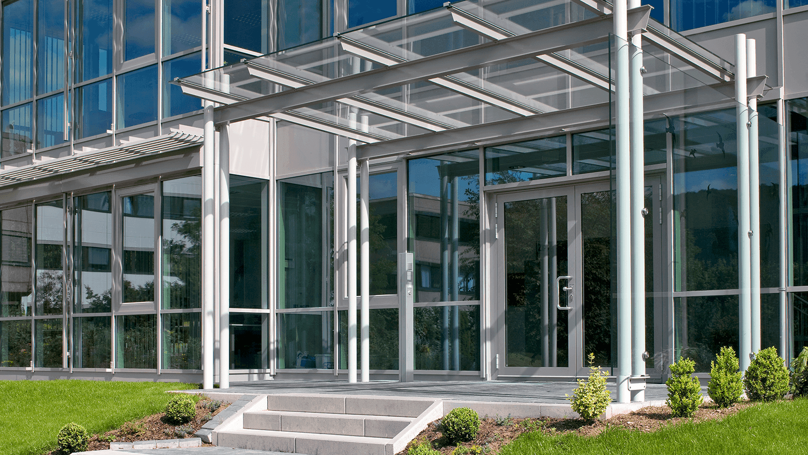 Moderner Glas- und Metallbau-Eingangsbereich mit Vordach und breiten Fensterflächen, umgeben von Grünflächen.