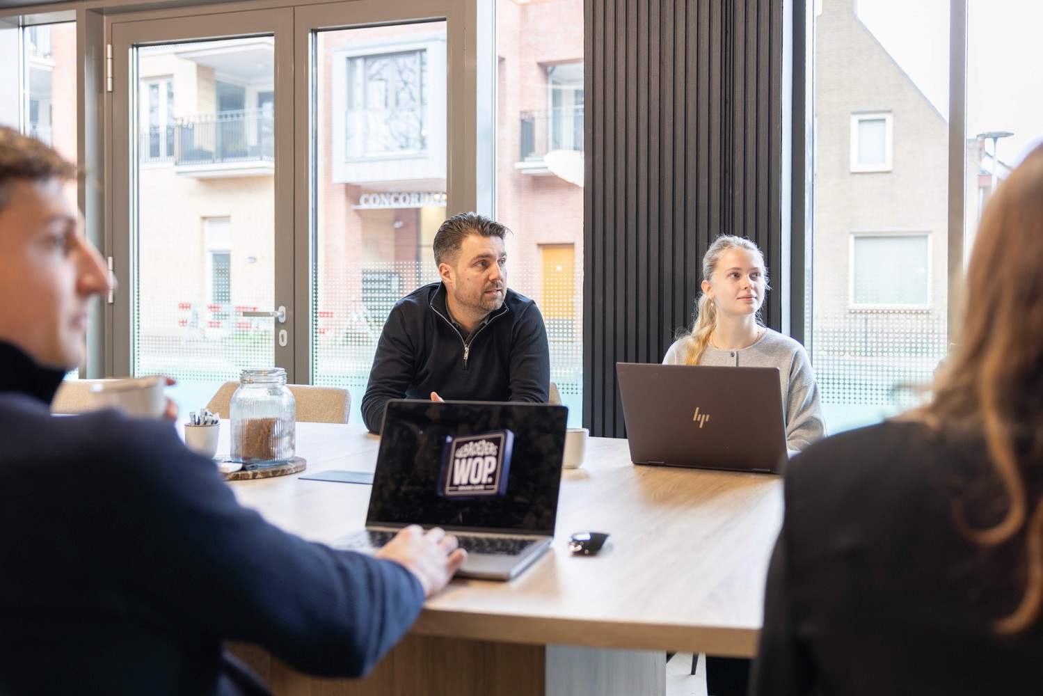 Close-up portrait of a person wearing a gray insulated hooded jacket with the hood up, showing their face framed by the hood against a light background.