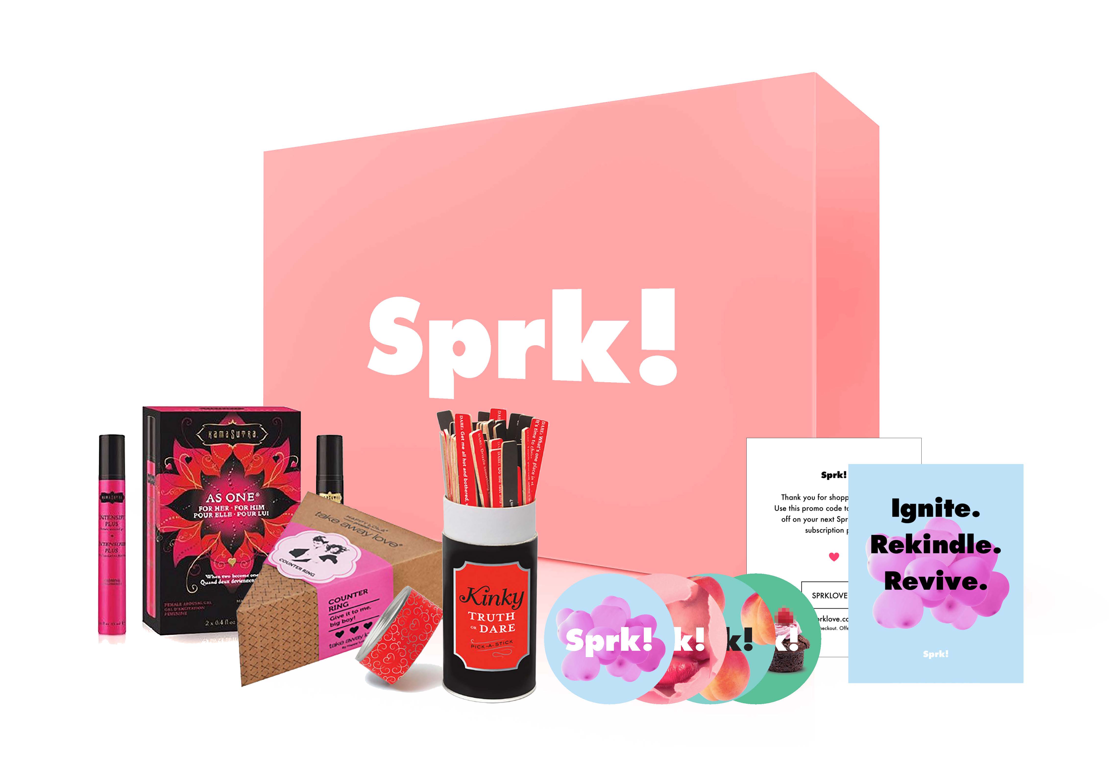 A pink box labeled “Sprk!” in white letters sits on a white background. In front of it are various items: a pink tube of lip gloss, a black box labeled “AS ONE” with floral designs, a black container labeled “Kinky Truth Dare,” a black container labeled “Kinky Truth,” a white card with a “Spark!” logo, a light blue card with a “Spark!” logo, a set of three colorful discs labeled “Sprk! K!,” and a light blue card labeled “Ignite. Rekindle. Revive.”.