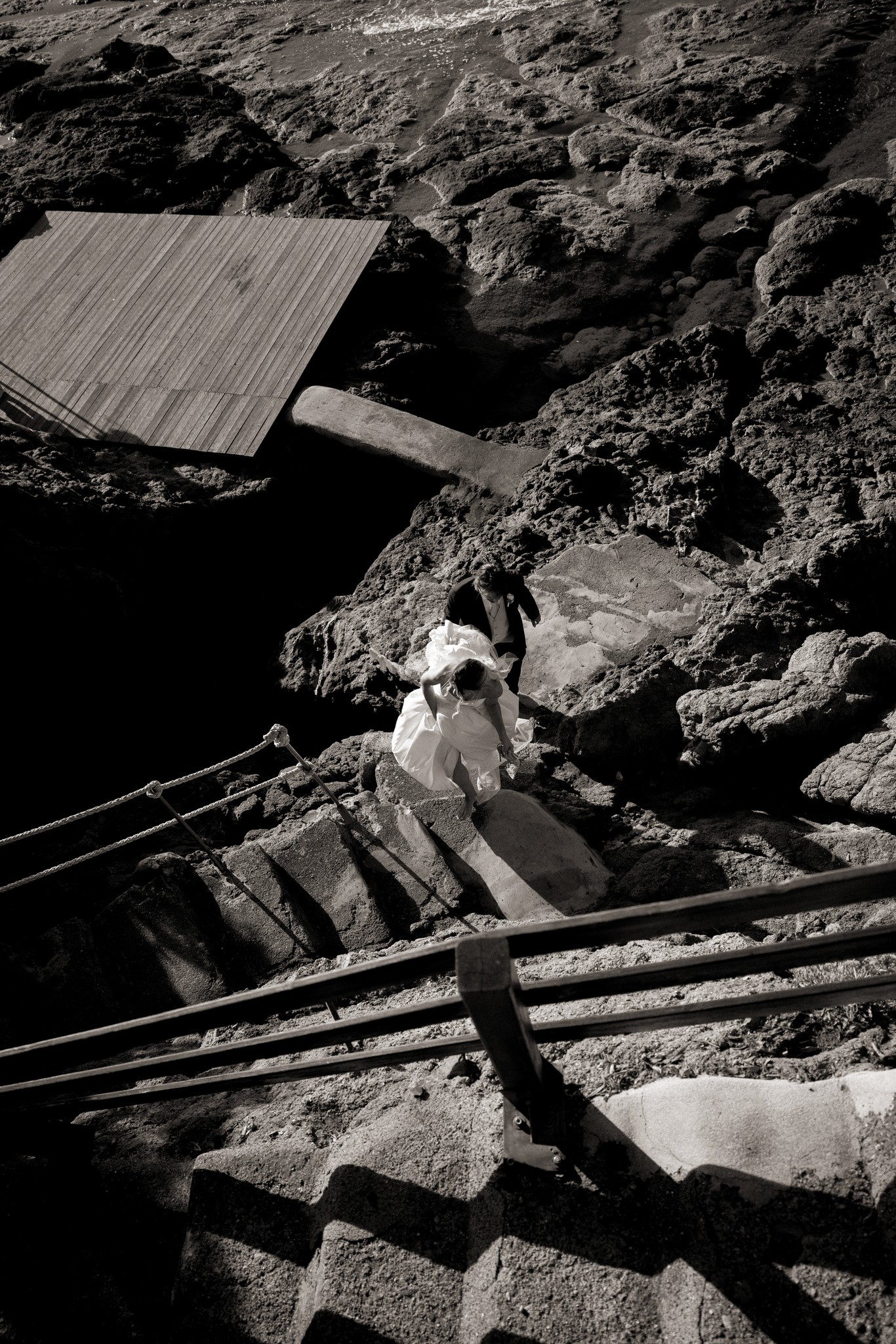 Sposi scendono verso la scogliera di Bolgheri, lui in abito scuro, lei in bianco, reportage matrimoniale in bianco e nero