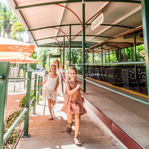 Three young girls joyfully run on a shaded walkway by a sign that reads "Endertom." The background includes greenery and empty benches.