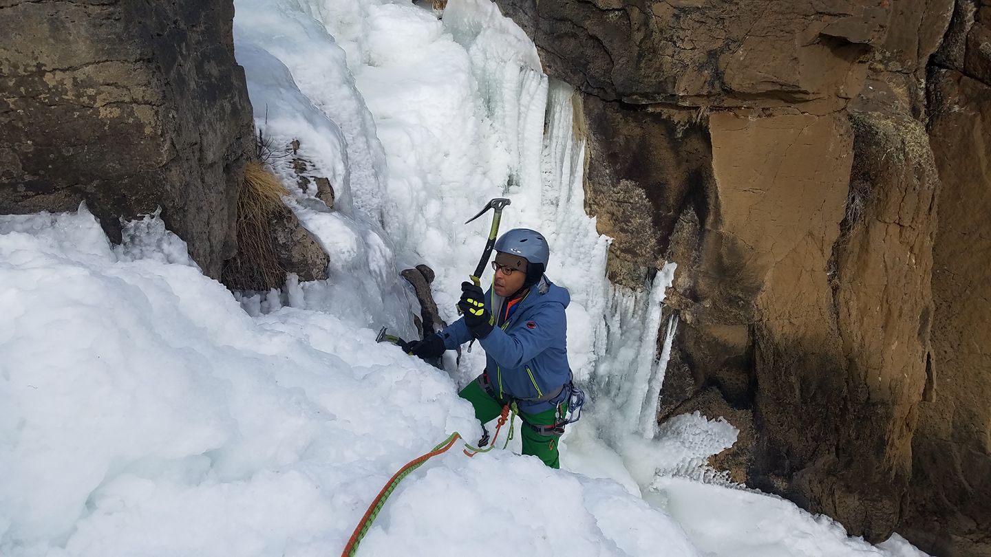 Ice climber in blue about to swing tool near top of frozen waterfall