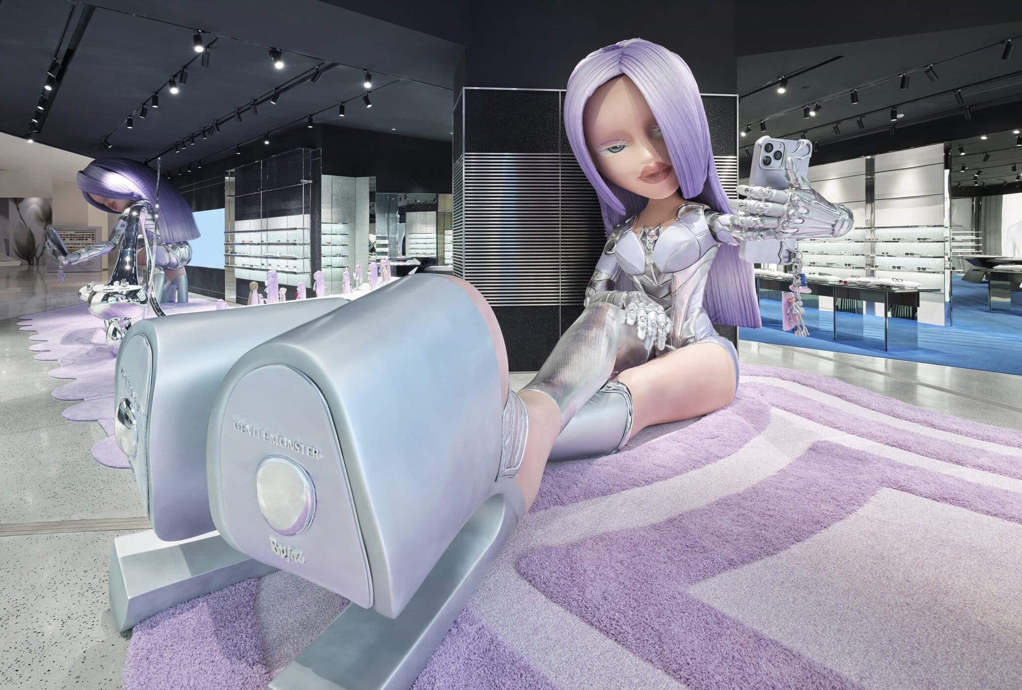 Inside a Gentle Monster store, a large Bratz-style doll with lavender hair reclines on a pastel display, wearing futuristic silver accessories amid a sleek retail interior.