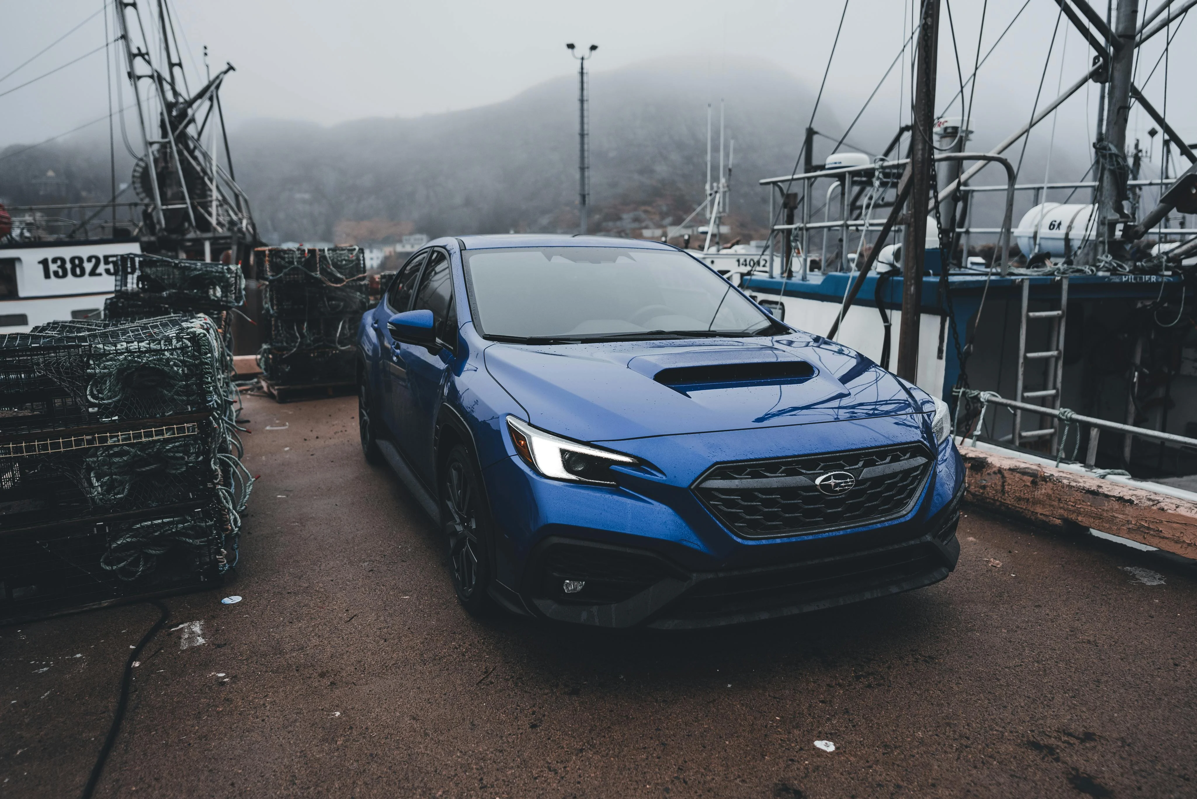 Subaru WRX bleue photographiée sur un port brumeux, entourée de filets de pêche, de bateaux et d’un décor marin industriel. La calandre agressive, la prise d’air sur le capot et la carrosserie mouillée mettent en valeur le caractère robuste et sportif du modèle, parfaitement à l’aise dans cet environnement humide et authentique.