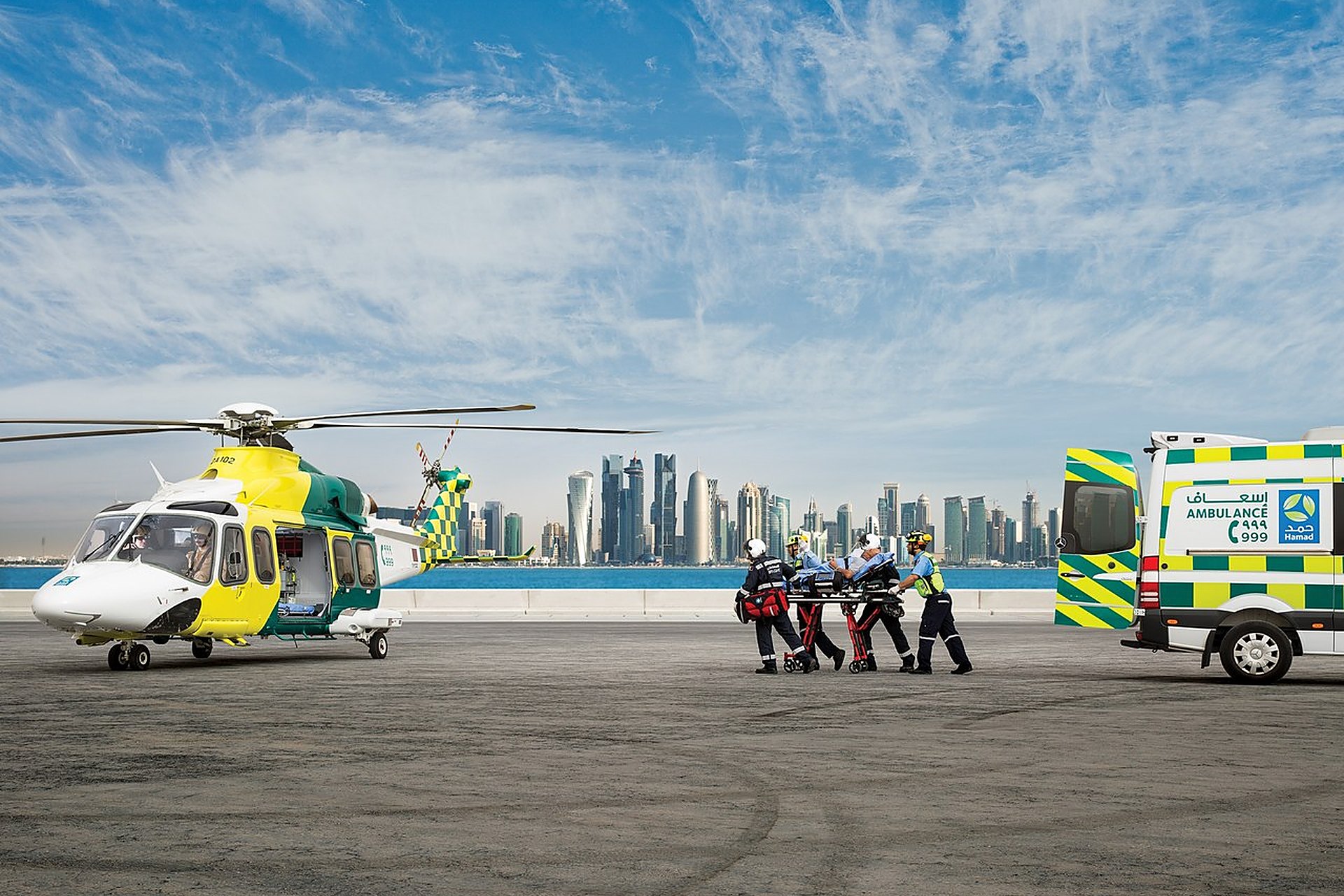 Un hélicoptère médicalisé jaune et vert posé face à la skyline de Doha, l'équipage en train de charger un brancard, une ambulance à côté
