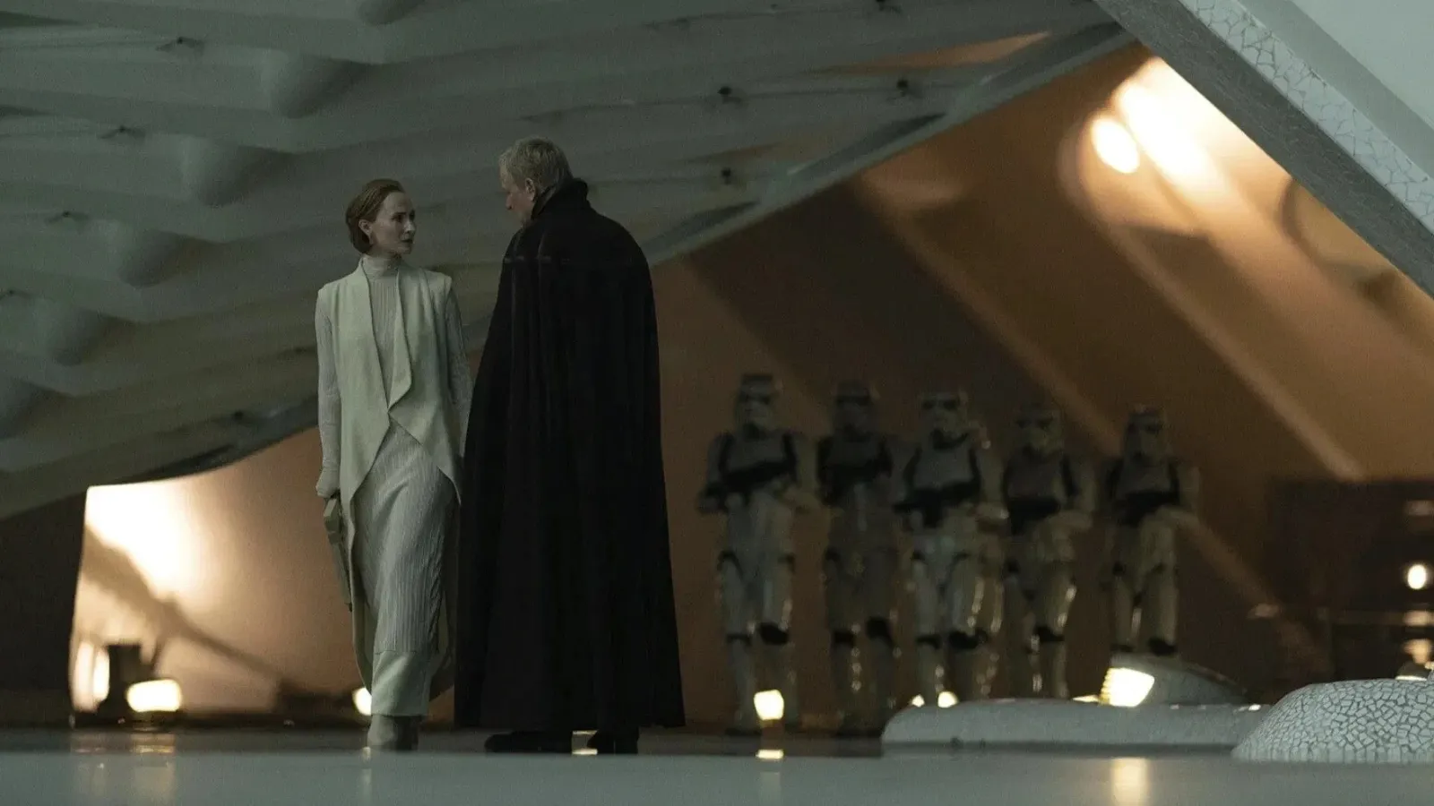 Under a futuristic, angular structure, two individuals in formal attire converse while a group of uniformed guards stands in the background, suggesting a tense, sci-fi setting.