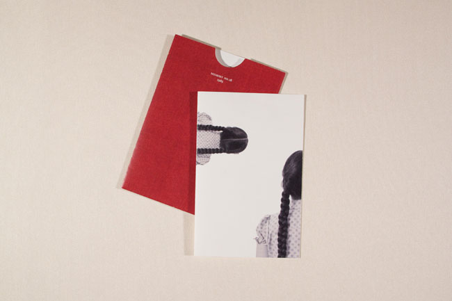 A red envelope lies flat on a light beige surface, partially covering a white card. The card displays a black and white photograph of a woman with long dark hair, viewed from behind, wearing a light-colored garment with a small dotted pattern. The woman’s head is turned to the right. The red envelope has white text on its top edge.