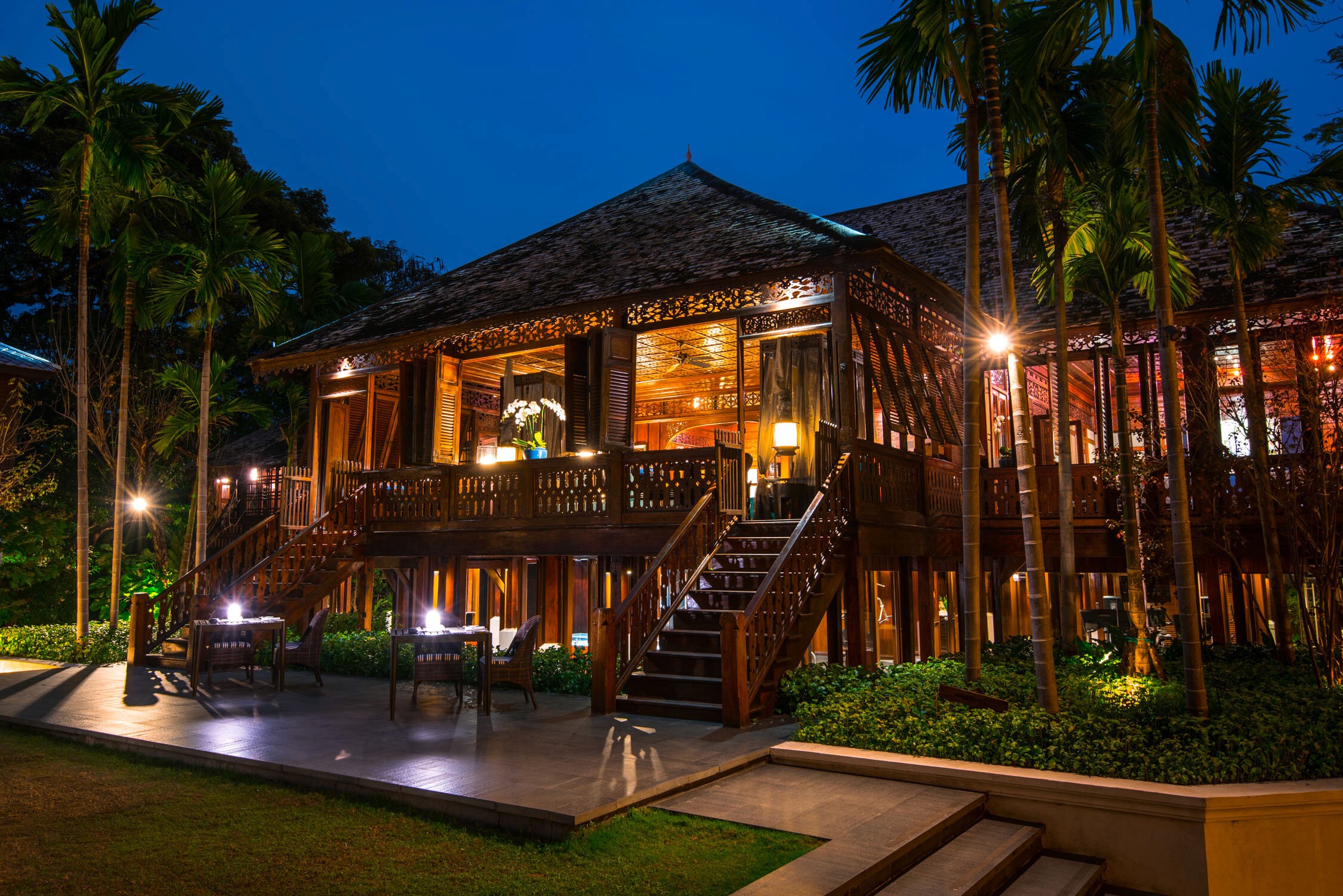A lit wooden resort villa with a balcony and stairs sits among palm trees under a dark blue twilight sky.