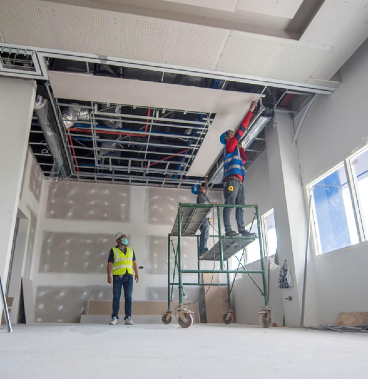 Two construction workers are at a job site; one is on a scaffold, while another stands on the ground observing.