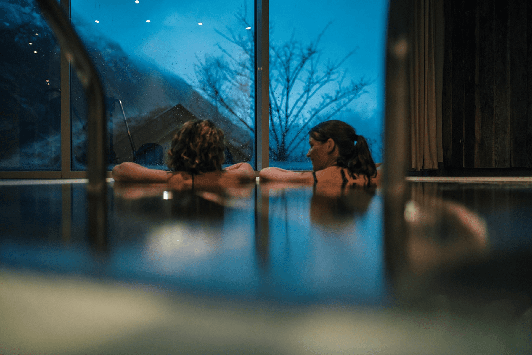 Two people relax in a serene indoor pool, surrounded by large windows showing a nighttime view.