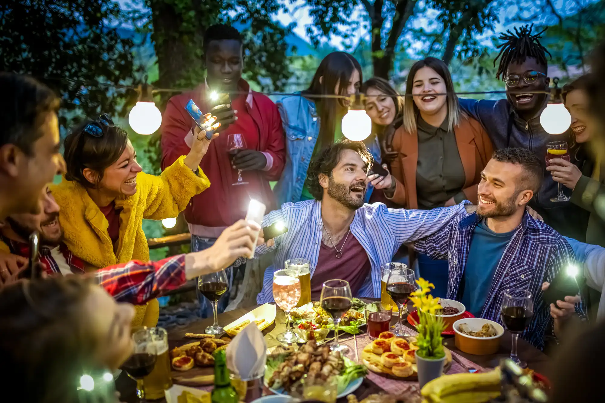 A group of young adults gatherd around a table outside full of food. 