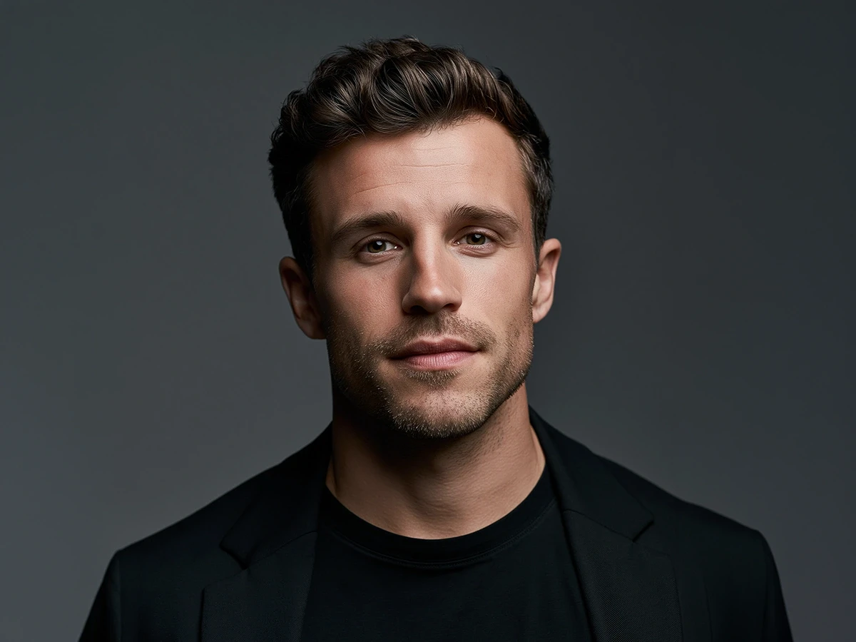 A studio headshot of a man with styled brown hair and light stubble, wearing a black jacket against a dark grey background.