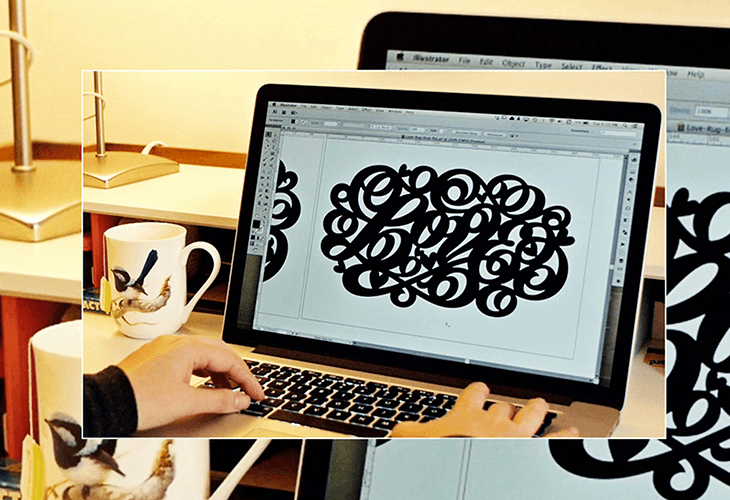 A hand types on a laptop keyboard with a black design on the screen. The laptop screen displays a black abstract design on a white background. To the left, a white mug with bird designs sits on a surface. A second laptop screen is partially visible behind the first. The background includes a light-colored wall and a lamp.