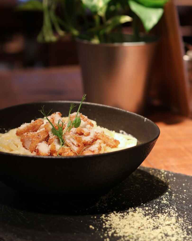A black bowl of creamy pasta topped with crispy bacon pieces, grated cheese, and fresh herbs on a wooden table.