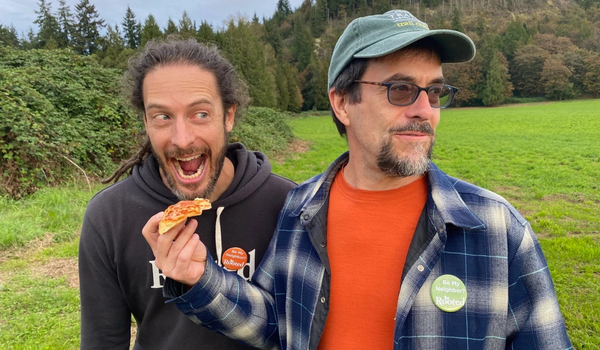 Two community members stand in a grassy field at Rooted Northwest, laughing as one enjoys a slice of pizza.