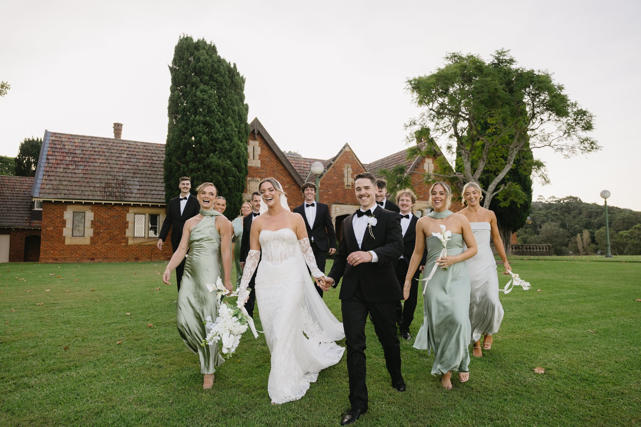Smiling bridal party in black tuxedos and sage dresses walk across a lawn before heritage brick buildings.