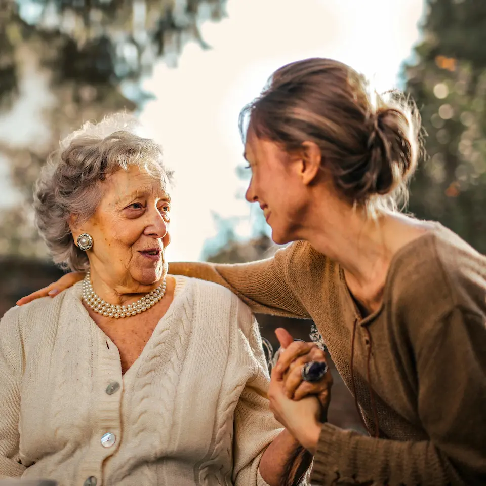 A younger woman leaning in warmly toward an older woman, holding her hand and sharing a gentle, connected moment together outdoors.