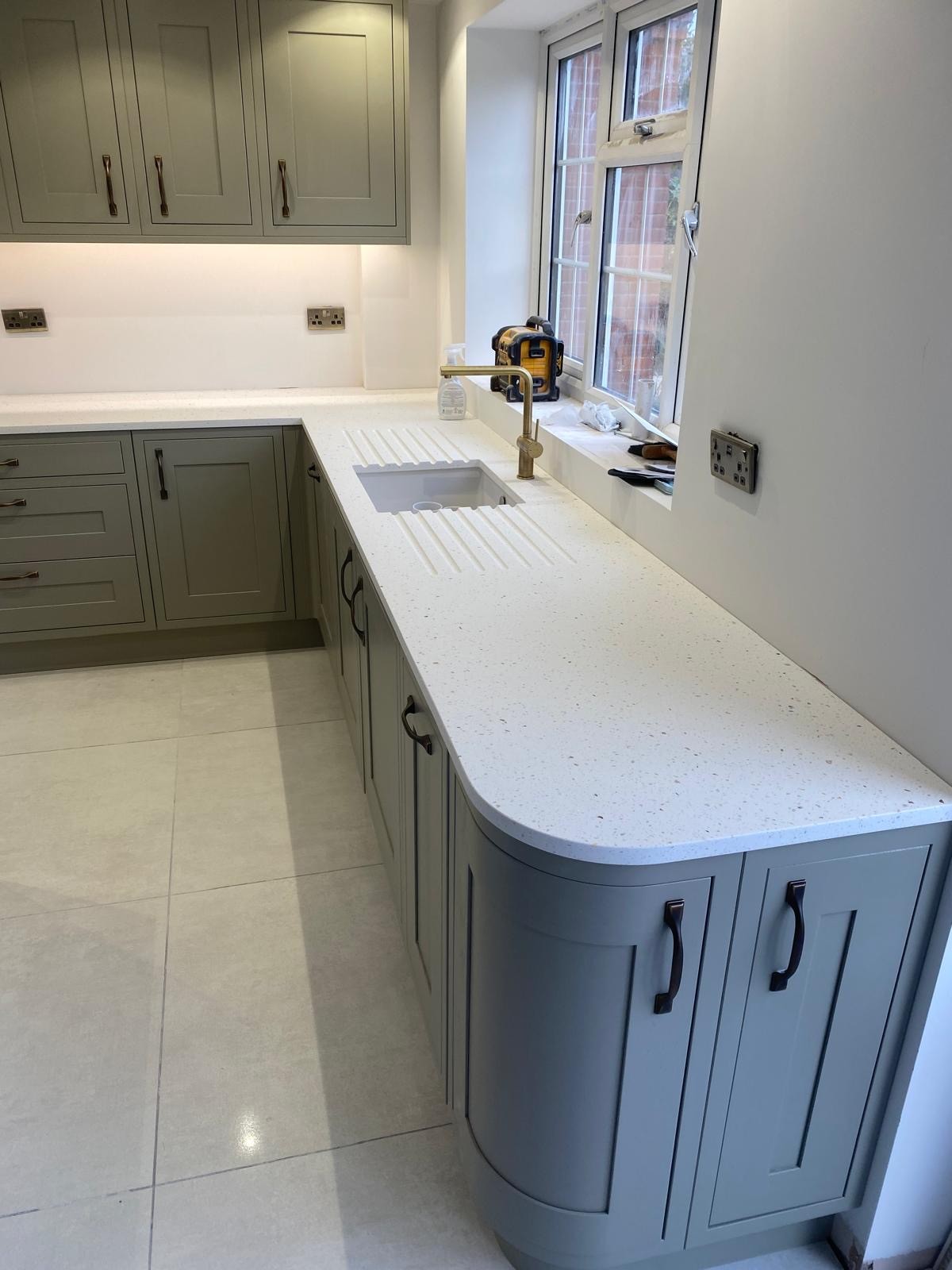 Bespoke sage green shaker kitchen with a curved quartz worktop and integrated sink, installed by PPM Builders in Northampton. Featuring elegant brass handles and under-cabinet lighting for a modern touch.