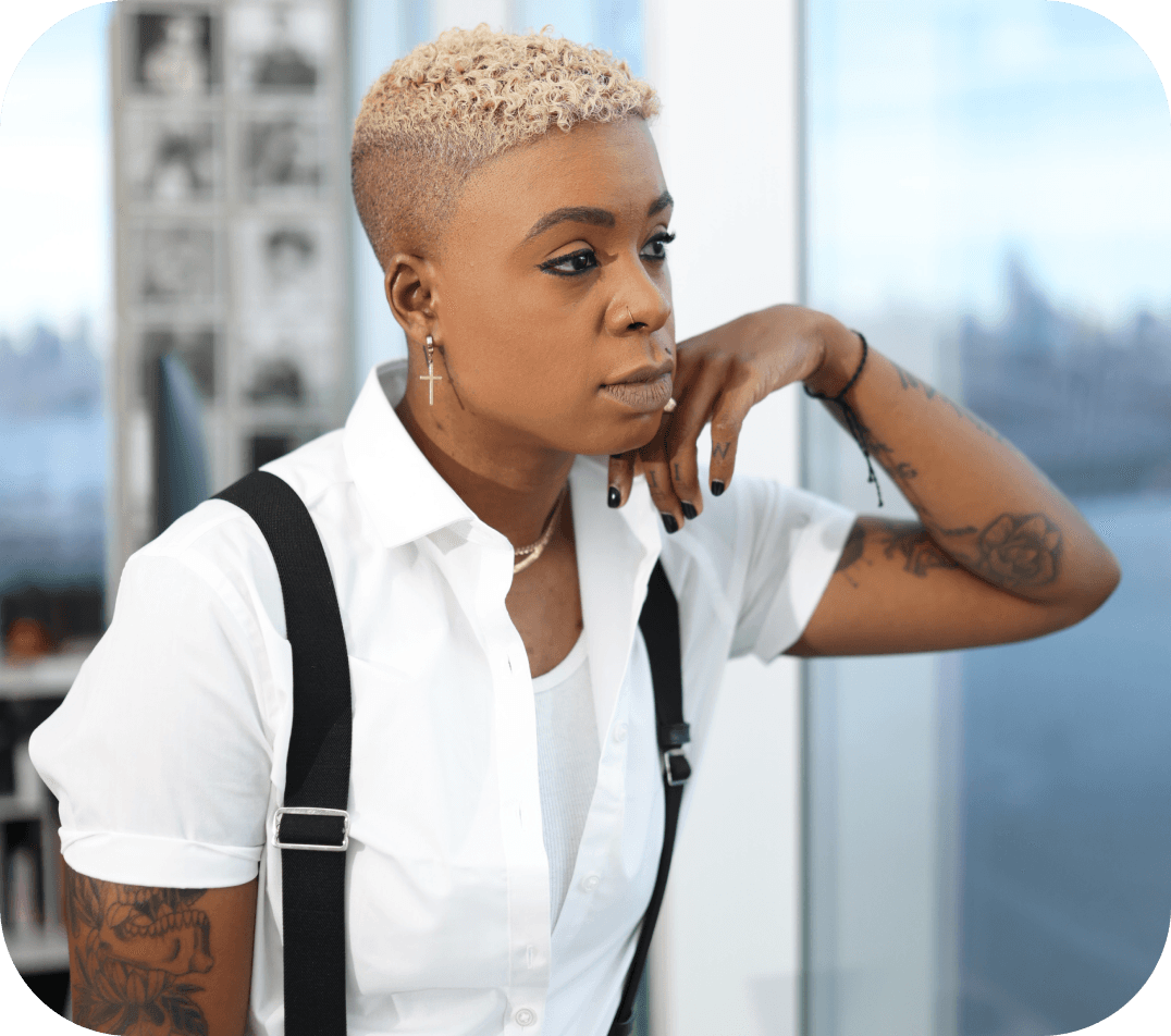Ashleigh Warren, a person with short blonde hair and tattoos wears a white shirt with suspenders, deep in thought against a city backdrop.