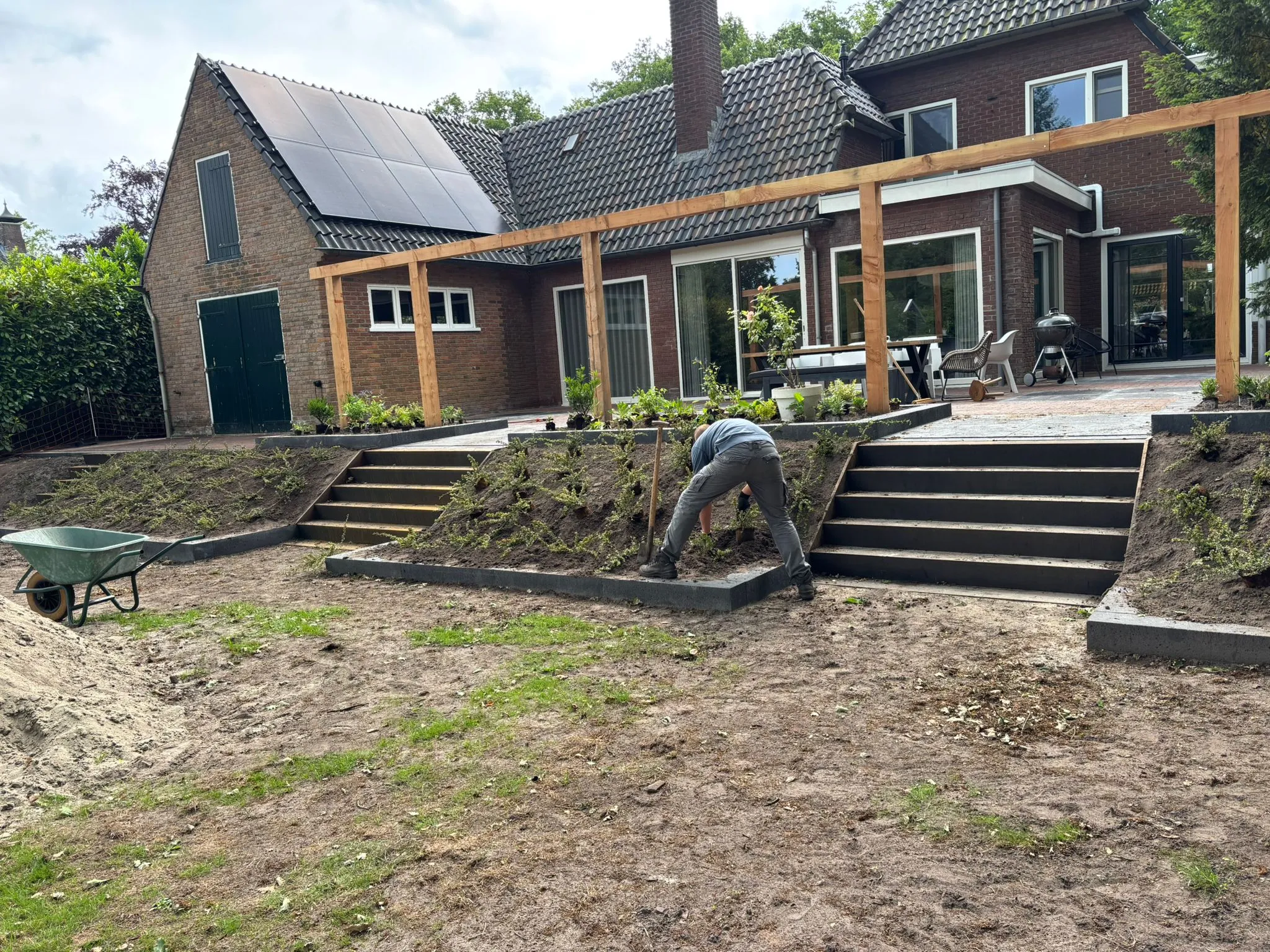 Rustiek terras met oud-Hollandse tegels en trap naar lager gelegen tuin, hoveniers planten struiken bij woning in Oldeberkoop.