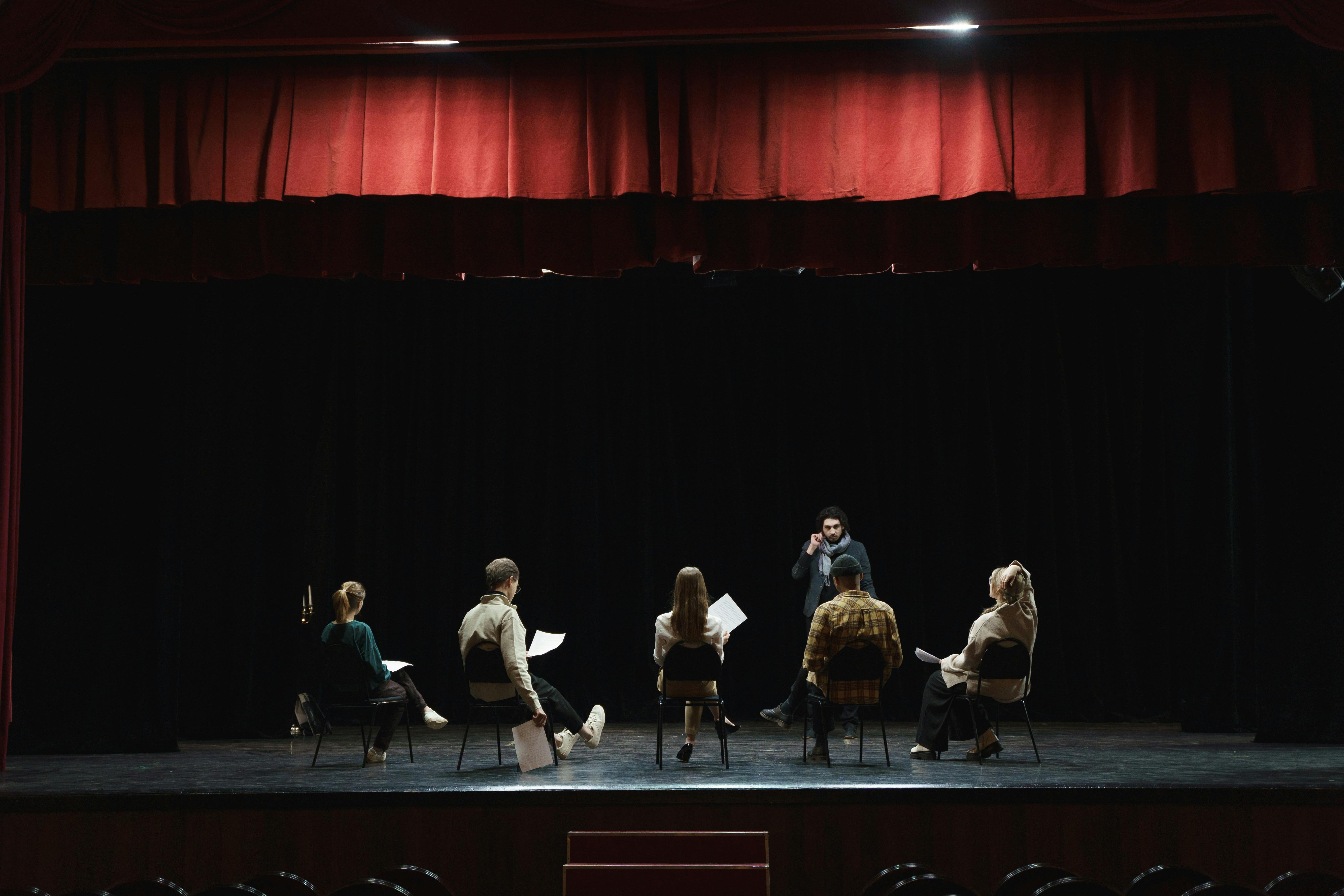 A group of actors sitting on stage with scripts in hand, facing a director giving notes during a theatre rehearsal under red curtains.