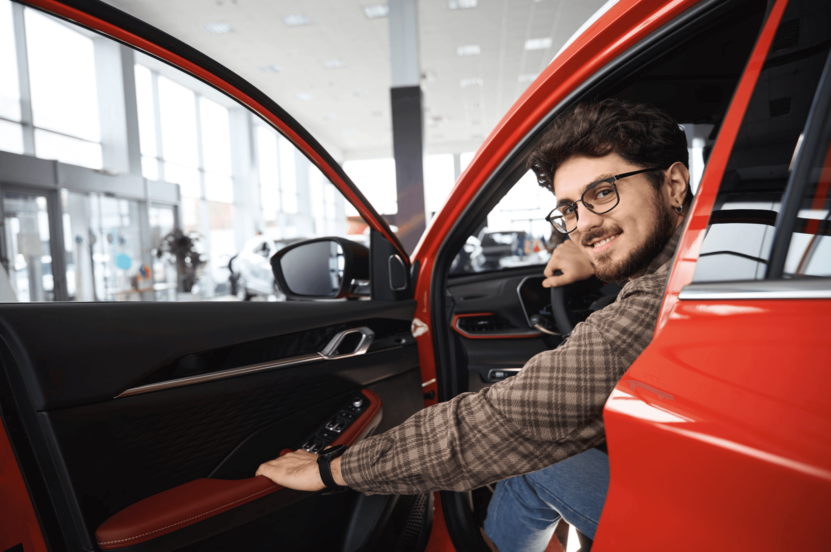 Young man sitting in the driver's seat of a red car inside a dealership, smiling confidently while testing out the vehicle.