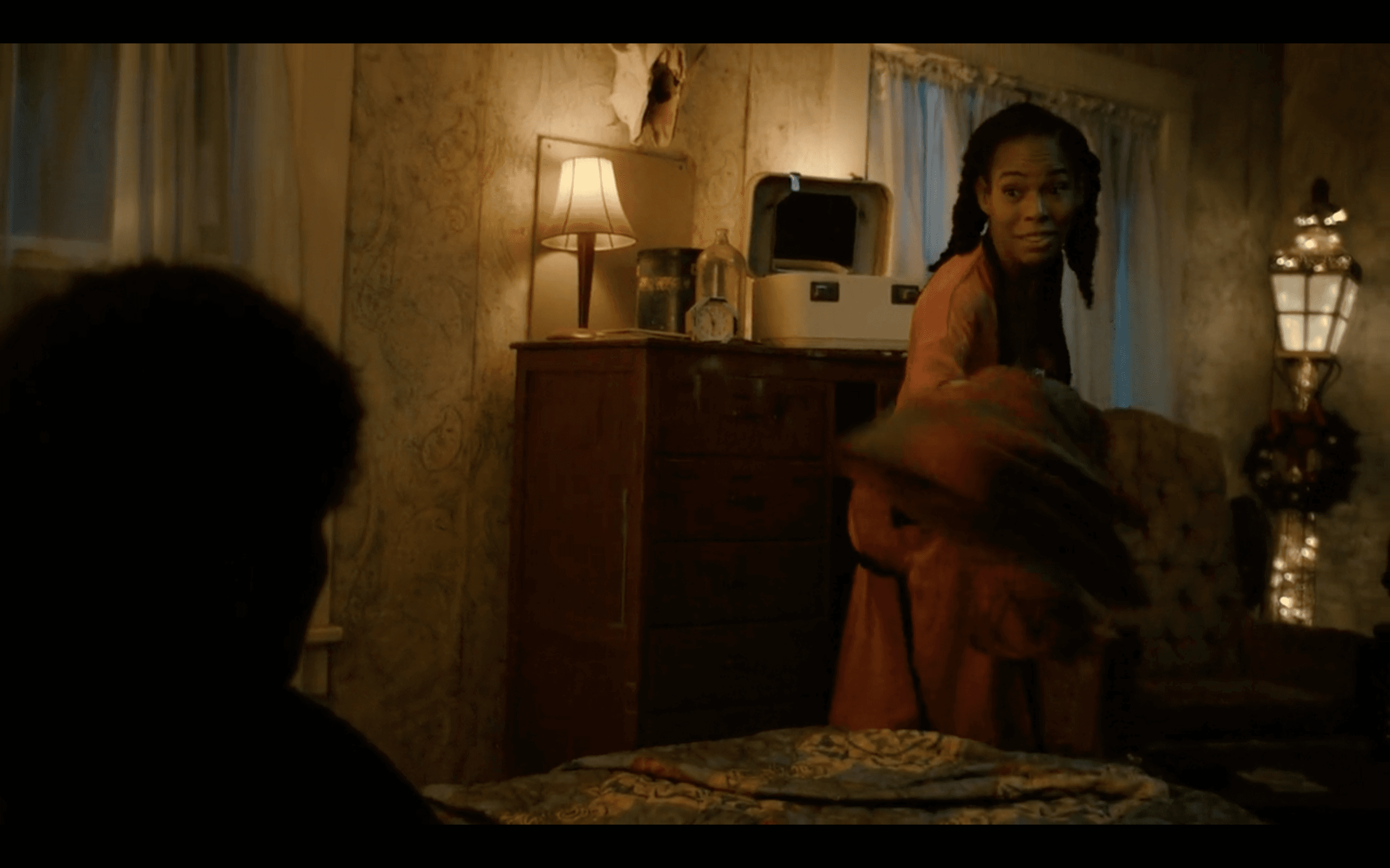 An African-American woman is talking to someone sitting at the corner of the frame, in a dimly lit house.