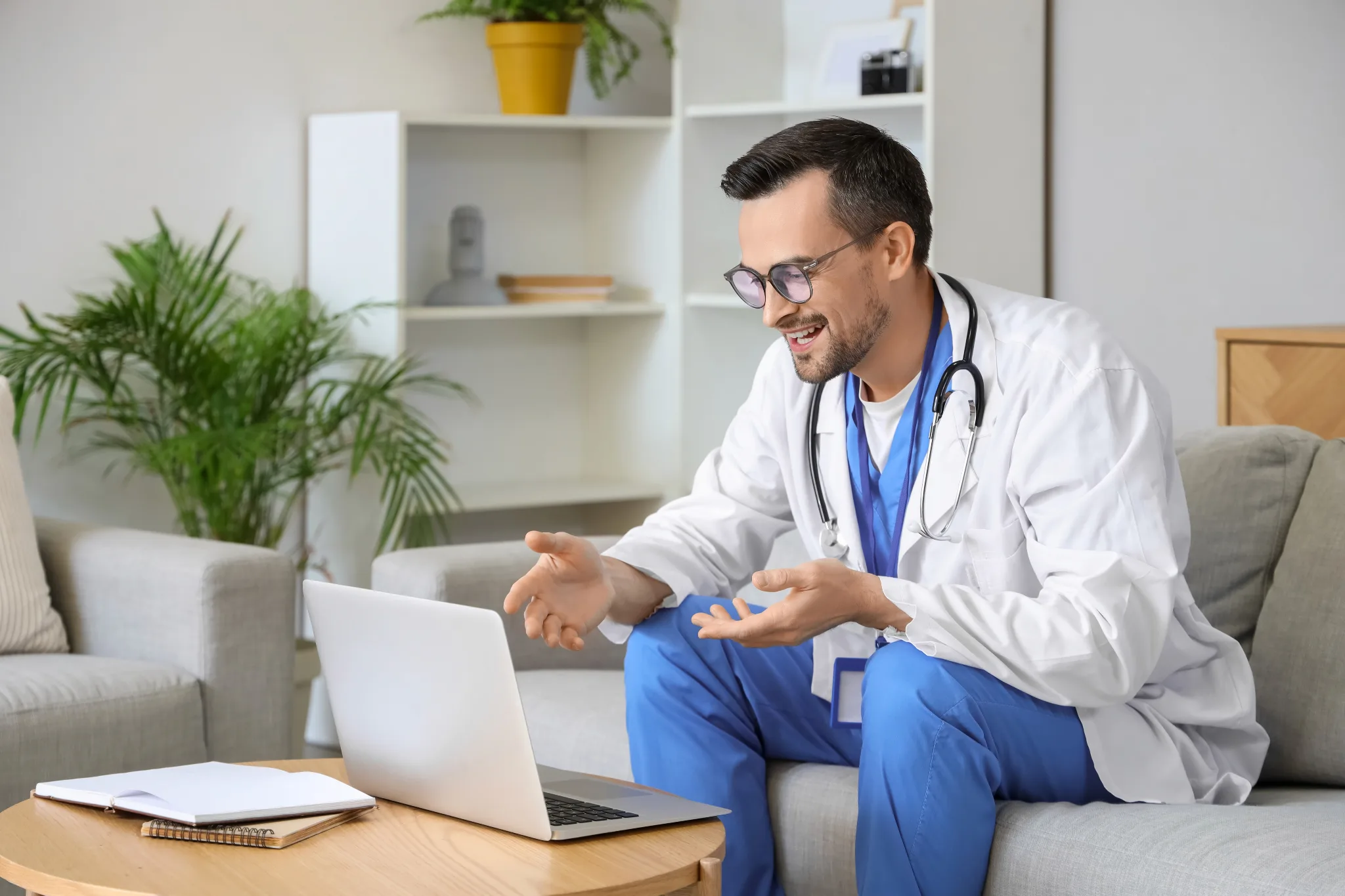 Male physician with glasses, wearing a white coat with stethoscope around the neck, sitting on a couch, laptop in front, talking to someone on the laptop.