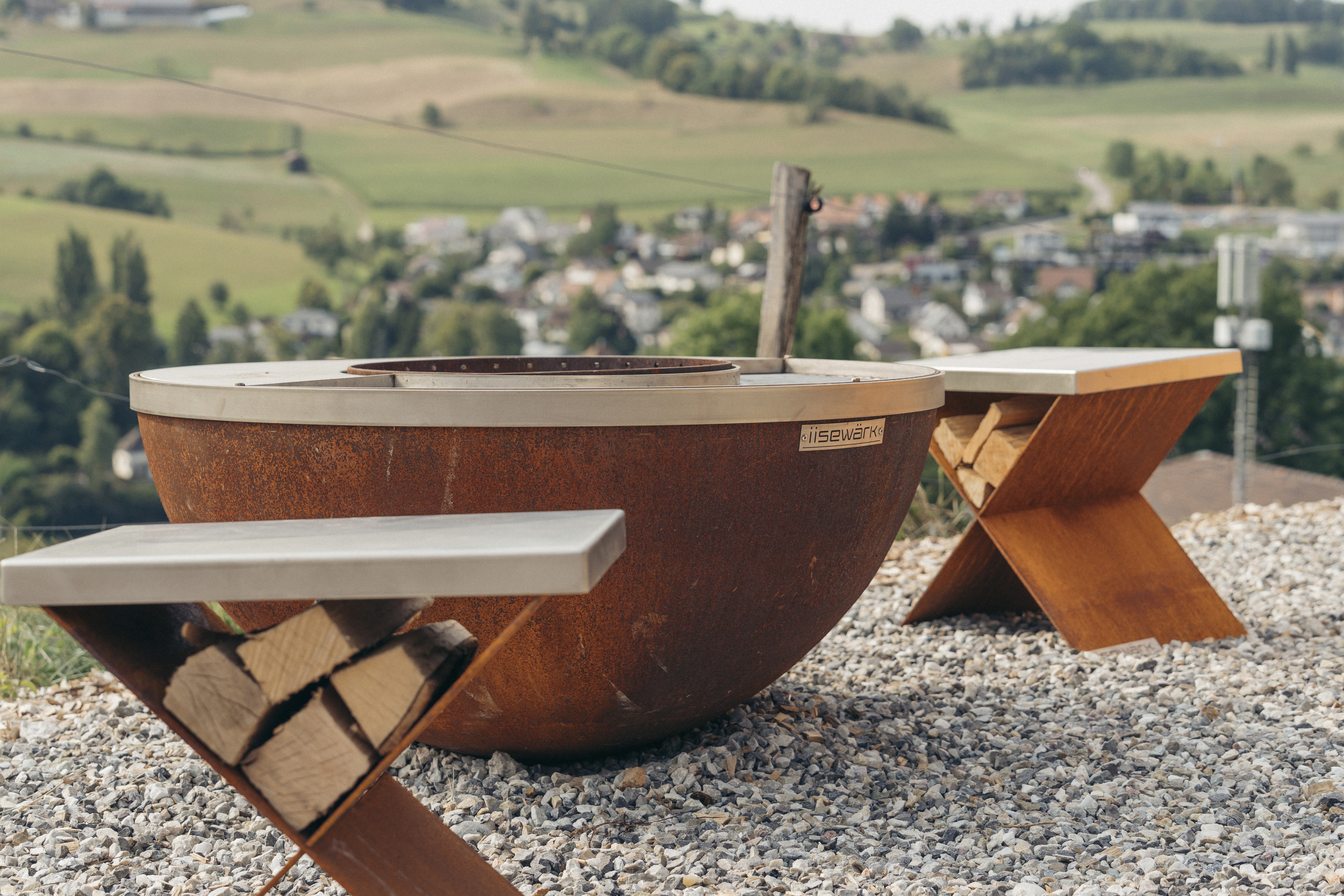 Eine großer, runder, rostfarbener, schalenförmiger Grill steht auf einem Kiesuntergrund, umgeben von Grünpflanzen und einer weitläufigen Landschaft.