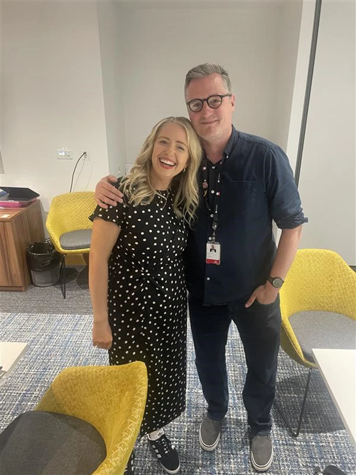 Two people standing close together in an office with yellow chairs and a table.