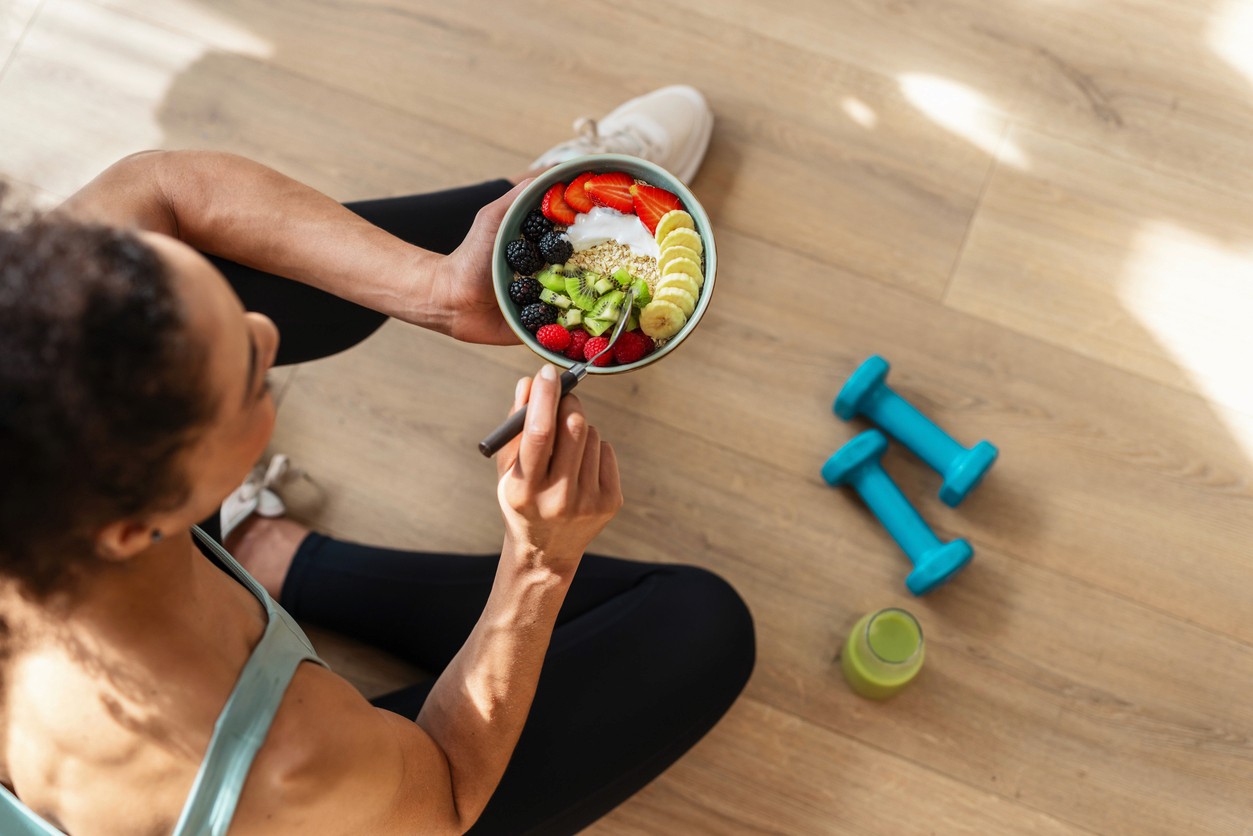 A women in exercise cothing sits on the floor eating a healthy meal after a working out with dumbells