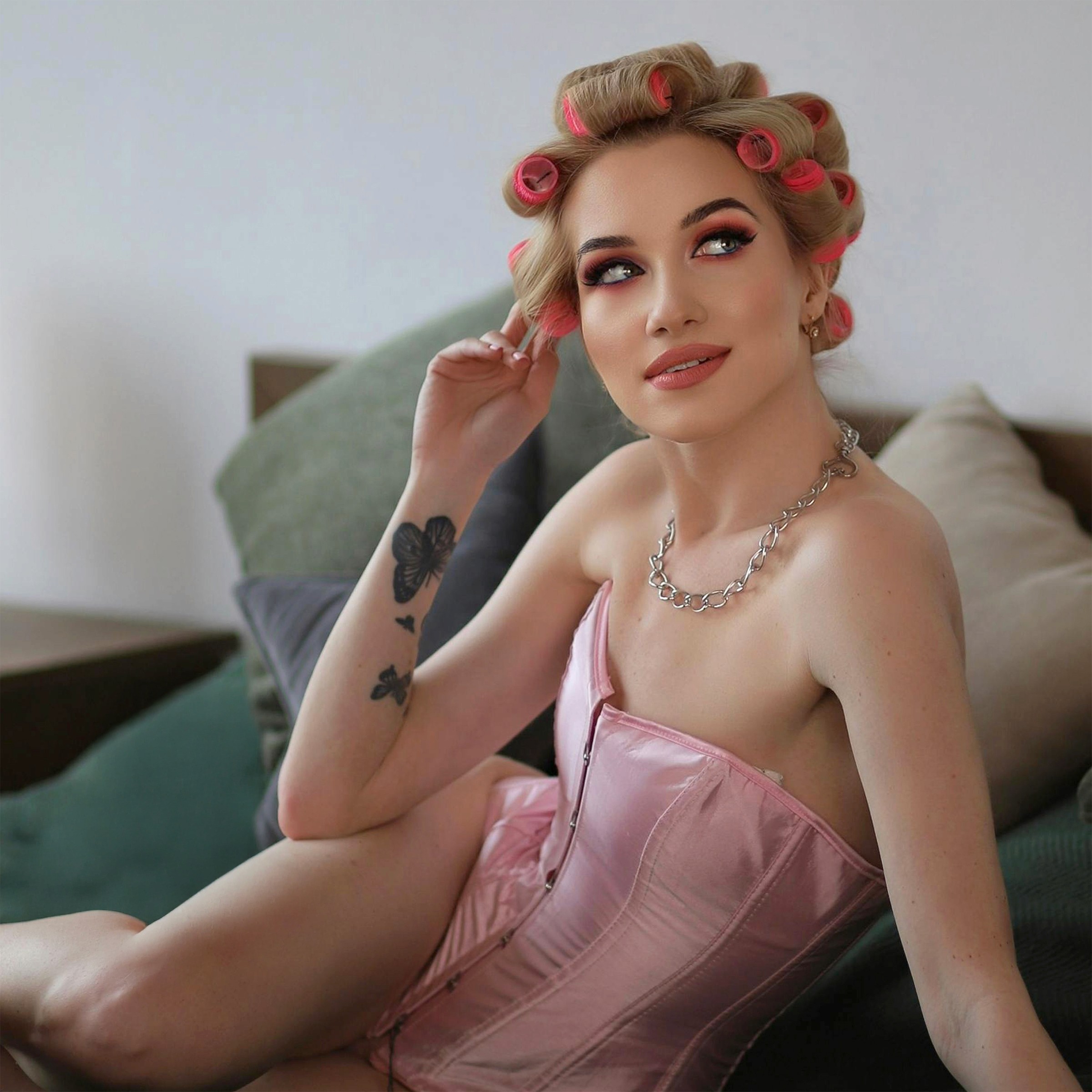 A blonde woman with pink hair rollers and a pink corset sitting indoors, displaying butterfly tattoos on her right arm.