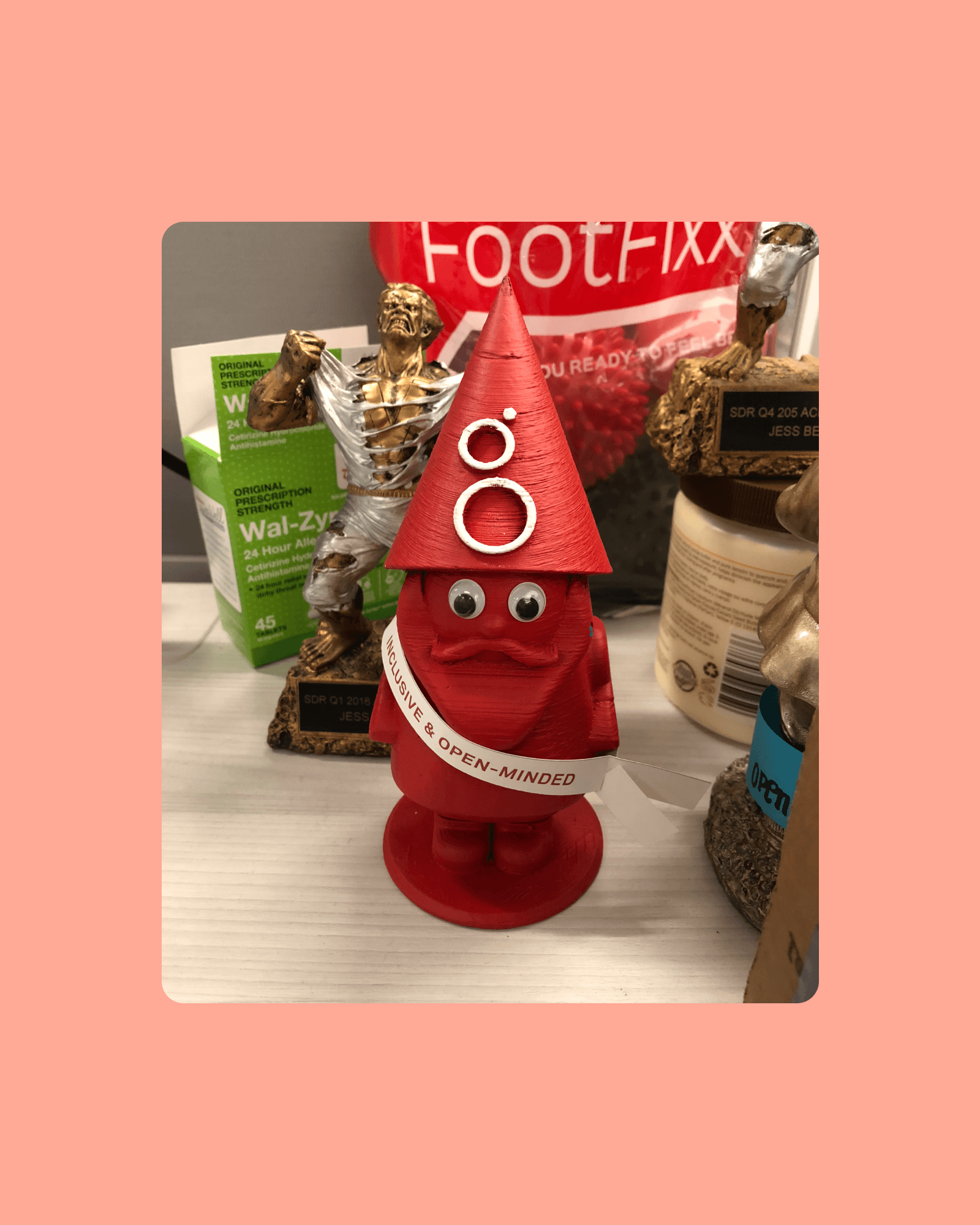 A red gnome sports googly eyes and a hand-labeled Inclusive and Open-Minded sash, proof coworkers personalized it as it traveled desks.