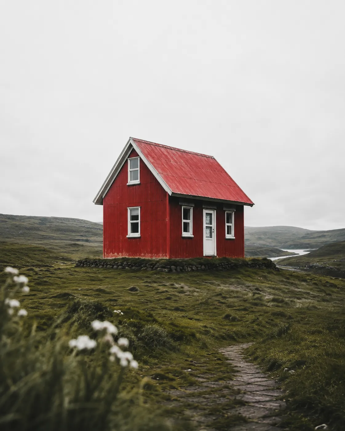 Red mountain chalet with a corrugated metal roof standing on a grassy hillside under an overcast sky, overlooking a winding river valley.