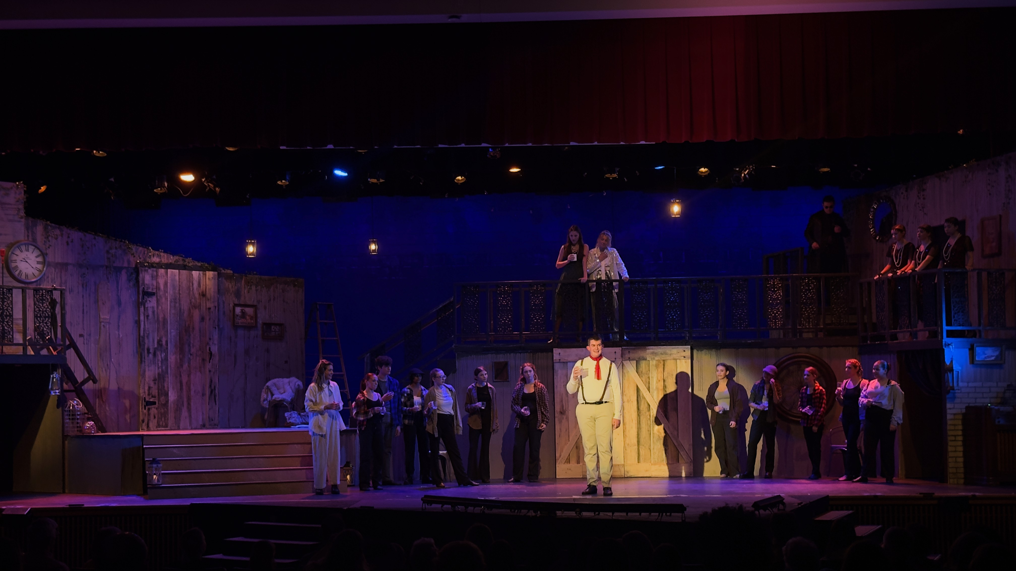 Orpheus stands center stage under focused lighting, surrounded by a dimly lit ensemble and a set washed in shades of purple and amber.