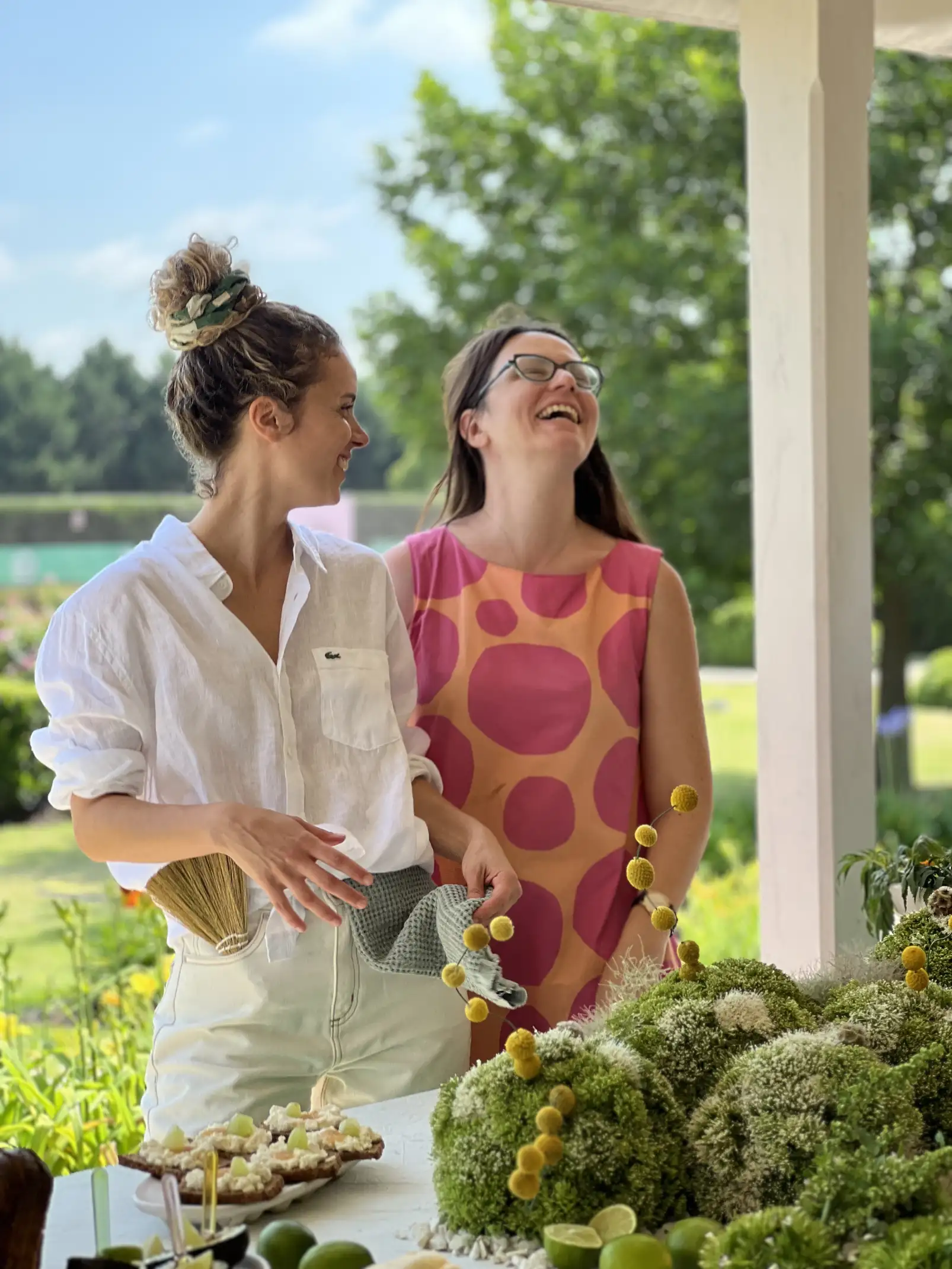 Dos mujeres sonríen y conversan al aire libre junto a una mesa decorada con arreglos florales verdes y amarillos, en un ambiente relajado y veraniego.
