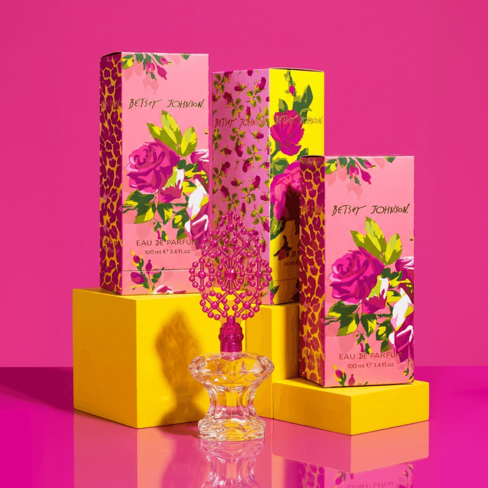 Betsey Johnson Parfum Produktinszenierung – kristallener Flakon mit pinker, perlenbesetzter Krone auf gelbem Sockel vor pinkfarbener Bühne, umgeben von vier gestaffelten Faltschachteln mit Rosen- und Leoparden-Prints.