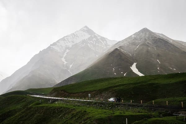 Mountain pass with green meadows, a road, and snowy peaks in the background.