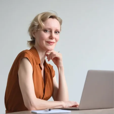 Woman with blonde hair in a low ponytail wearing a terracotta sleeveless blouse, sitting at a desk with hand on chin, looking at camera with laptop in front of her