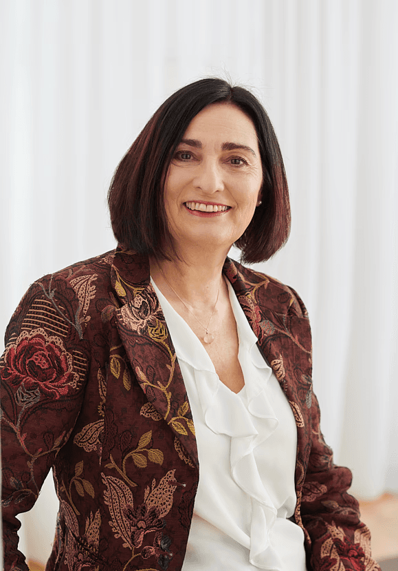 Dr. med. univ. Ursula Pueringer, MSc, systemischer Coach, Ärztin für Allgemein- und Arbeitsmedizin und Betriebliche Gesundheitsförderung trägt ein rotes Blumensakko und steht vor einem weißen Vorhang.