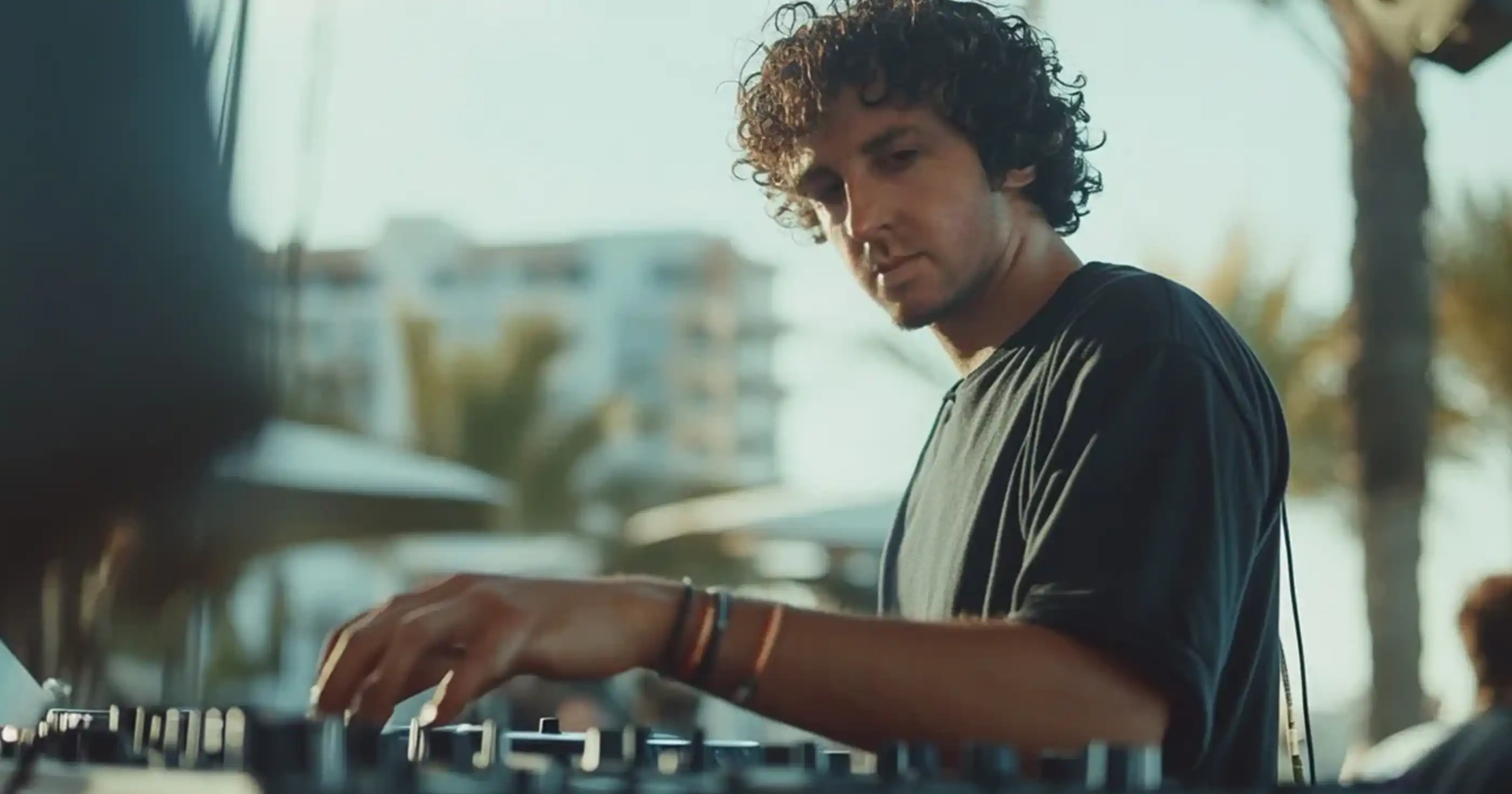 Jamie XX performing at an outdoor event, adjusting mixer controls with a focused expression at sunset.