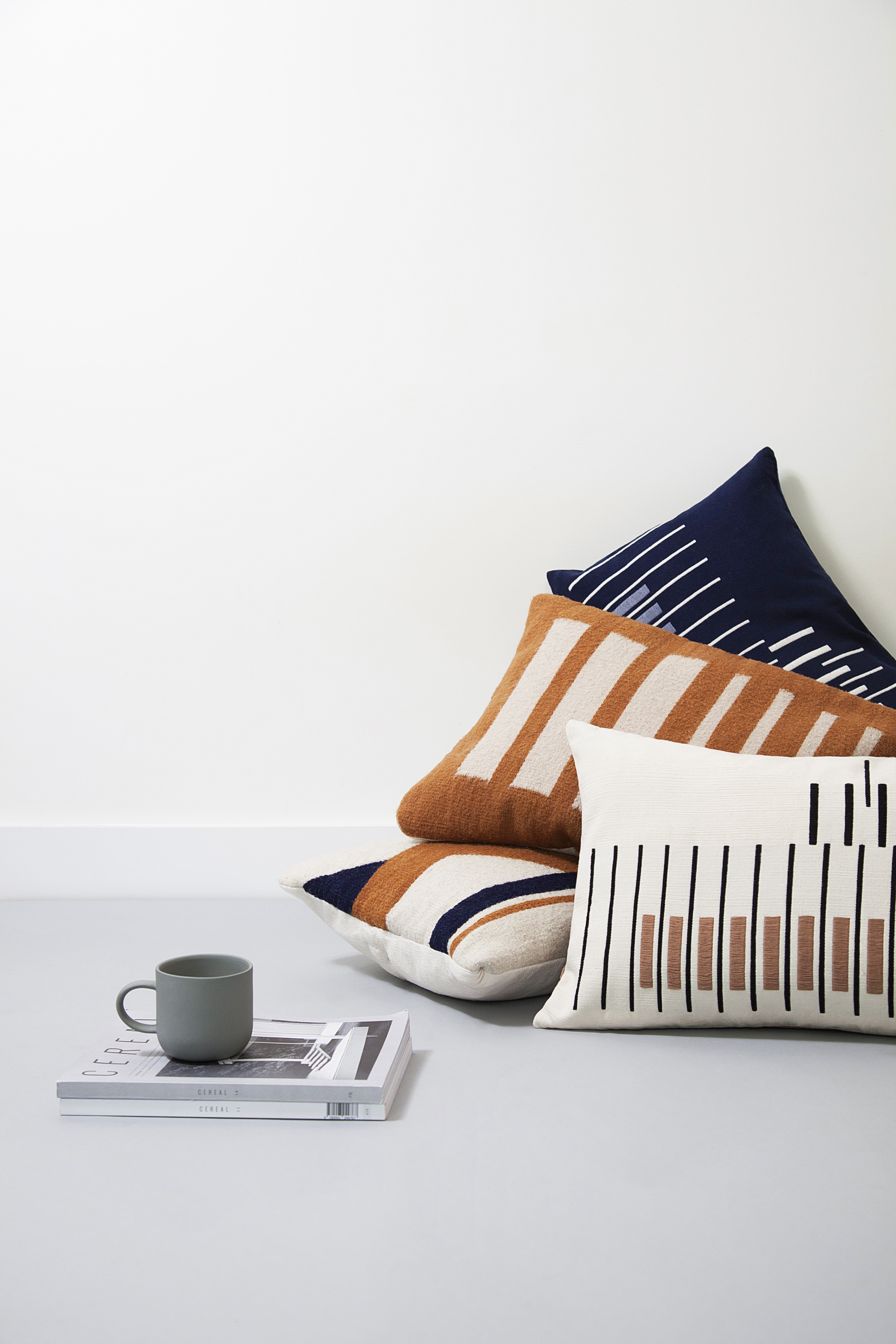 Three decorative pillows with geometric patterns in earthy tones are stacked against a minimalist white wall, next to a small stack of magazines and a grey ceramic mug on a light grey floor.