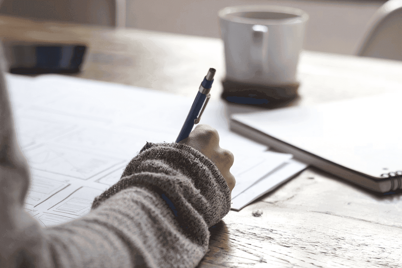 Person writing notes by hand at a wooden desk with coffee nearby.