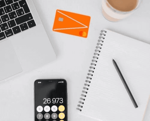 하얀 책상 위에 놓인 실버 노트북(맥북 맥락) 일부, 주황색 신용카드 한 장, 계산기 앱이 켜진 스마트폰(화면에 26,973 숫자), 그리고 볼펜 한 자루와 줄 노트를 위에서 내려다본 항공샷 사진입니다. 알뜰폰 요금제 비교 중임을 상징합니다.