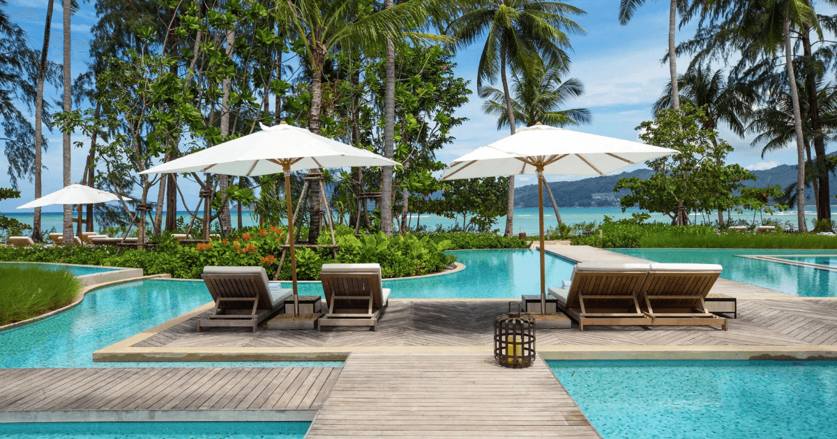 A resort pool with sun loungers and umbrellas surrounded by palm trees overlooking the ocean.